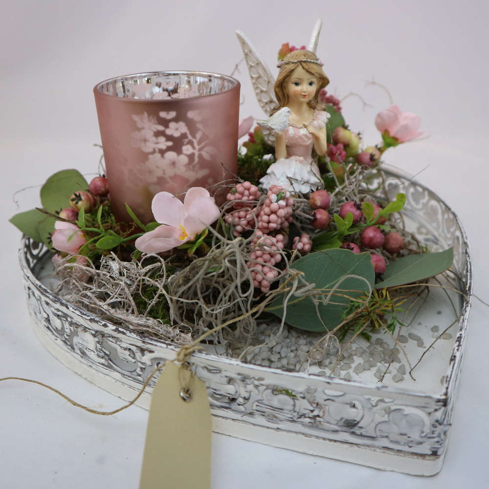  Zwischen Zweigen und rosa Beeren, flankiert von einem Kerzenglas, sitzt eine kleine Elfe in einem märchenhaften, rosa Kleid. 