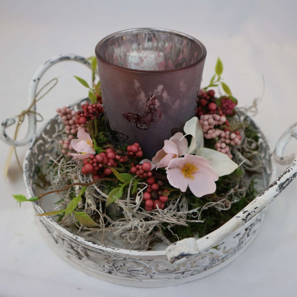 Ein weißes Tablett, dekoriert mit einem Kerzenglas und Kunstblumen sowie -beeren.