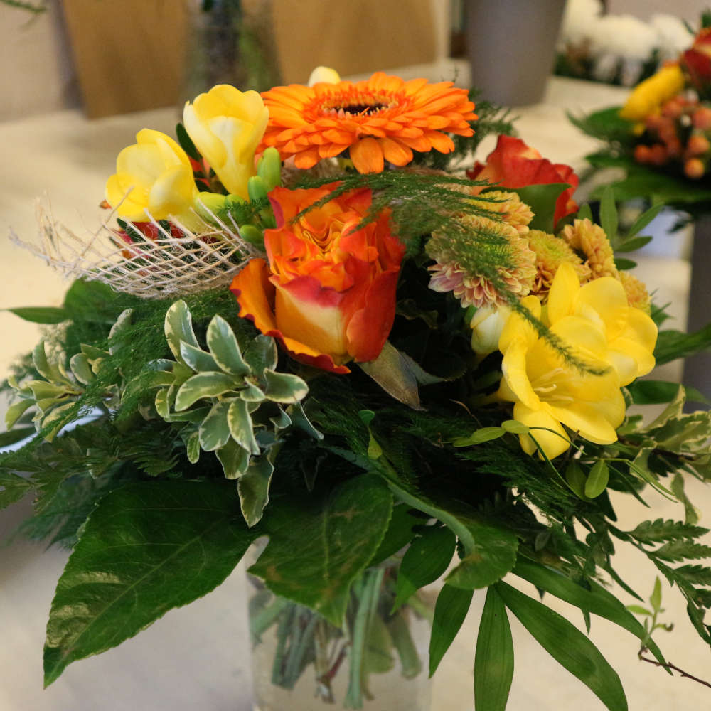 Ein kleiner, kompakt gebundener Blumenstrauß, bestehend aus orangen Gerbera und orange-gelben Rosenblüten; ergänzt um gelbe Blüten.