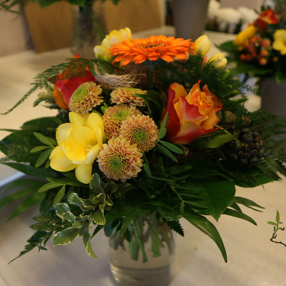 Ein kleiner, kompakt gebundener Blumenstrauß, bestehend aus orangen Gerbera und orange-gelben Rosenblüten; ergänzt um gelbe Blüten.